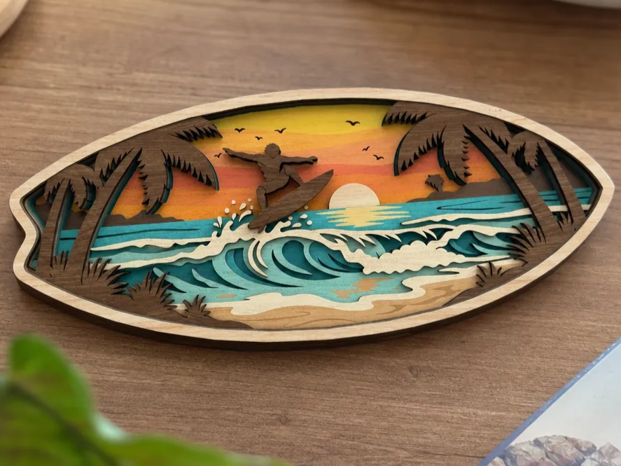 Layered wall art piece featuring a surfboard, a sunset over a tropical beach, and surfers