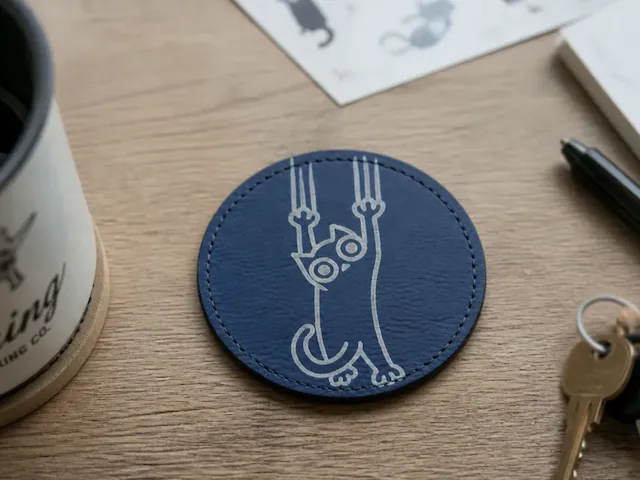 Blue circular leather coaster featuring a playful cat illustration