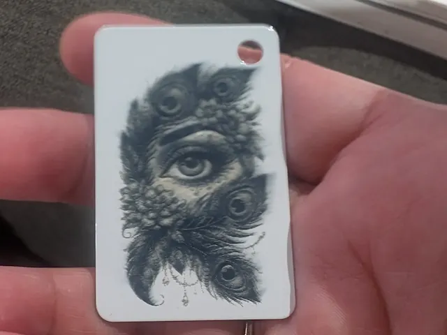 Rectangular keychain tag featuring an eye and peacock feather illustration