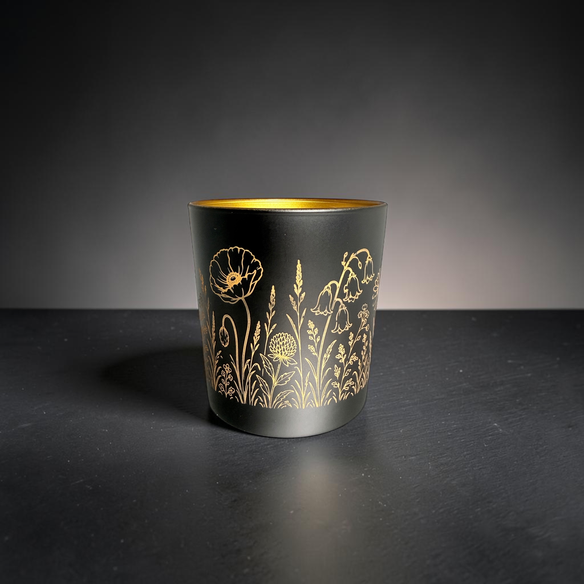 [Laser cut and engrave] Black candle holder with gold interior, engraved all around with a delicate wildflower meadow design using a rotary laser setup.
