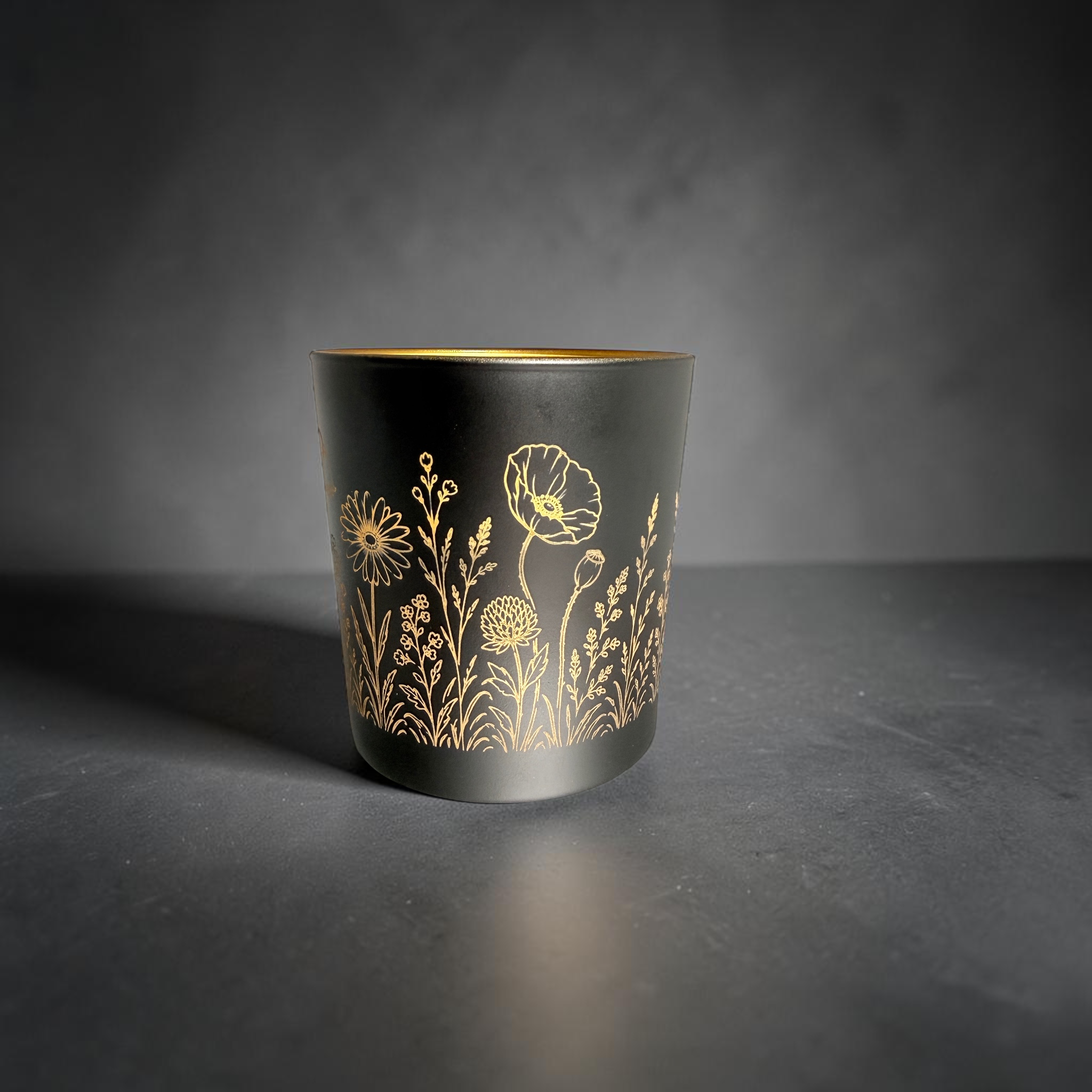 [Laser cut and engrave] Black candle holder with gold interior, engraved all around with a delicate wildflower meadow design using a rotary laser setup.